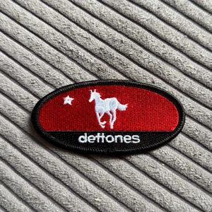 May include: An oval patch with a red background and a white horse silhouette. The patch has a black border and the word "deftones" in white. A white star is in the upper left corner.