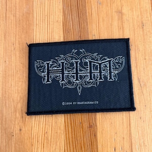 May include: Black rectangular patch featuring the word "HIM" in white, decorative lettering. The text "©2004 OY HEARTAGRAM LTD" is below. The patch is on a wooden surface.