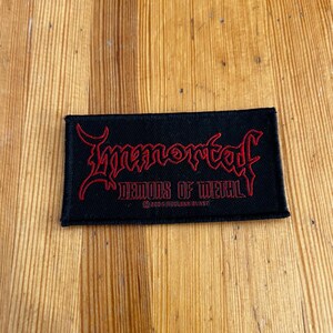 Könnte beinhalten: Schwarzer rechteckiger Aufnäher mit roter gotischer Schrift, die „Immortal“ über „Demons of Metal“ lautet. Der Aufnäher enthält auch den Text „©2006 NUCLEAR BLAST“. Der Aufnäher befindet sich auf einer Holzoberfläche.