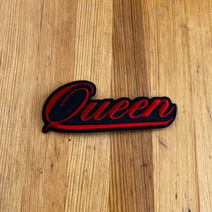 Op de afbeelding: Een zwart embleem met het woord "Queen" in rode letters geborduurd. De patch heeft een rode rand en ligt op een houten achtergrond.