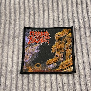 Puede incluir: Un parche cuadrado negro con un diseño detallado y colorido. El parche presenta el texto "MORBID ANGEL" en rojo y amarillo, junto con una ilustración de una estructura gótica y un fondo oscuro y ominoso.