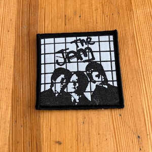 Op de afbeelding: Vierkante zwart-wit patch met een bandillustratie. De patch heeft een rasterachtergrond met de bandnaam "The Jam" in een gestileerd lettertype. Onder de tekst staan drie silhouetten.