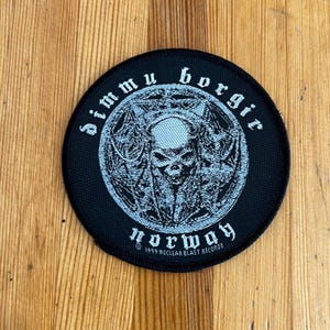 Puede incluir: Un parche bordado en blanco y negro con el texto "Dimmu Borgir Norway" y un diseño de calavera. También se incluye el texto "1999 Nuclear Blast Records".