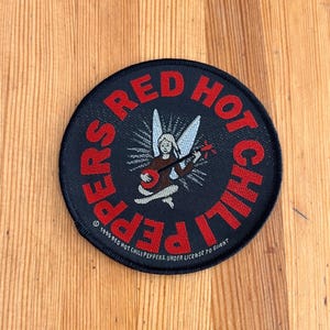 May include: A round, black patch featuring the text "RED HOT CHILI PEPPERS" in red. The patch includes an illustration of an angel playing a guitar. The text "© 1995 RED HOT CHILI PEPPERS. UNDER LICENSE TO GIANT" is also present.