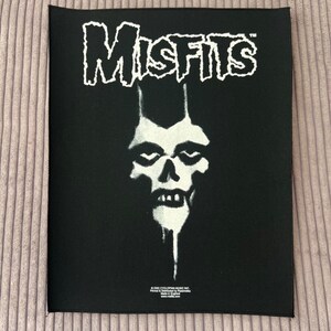 May include: Black patch featuring the band name "Misfits" in white, stylized lettering above a white skull graphic. The patch is rectangular and has a black border. The image includes text at the bottom.