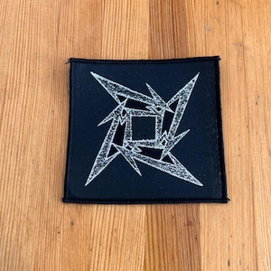May include: Black square patch with a white, stylized, four-pointed star design. The patch has a black border and is set against a wooden background. The design is a geometric representation of a star.
