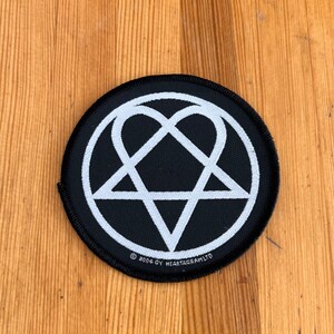 May include: A circular black patch with a white heartagram design, a heart-shaped symbol within a pentagram. The patch has a black border and the text "© 2004 OY HEARTAGRAM LTD" is printed along the bottom edge. It rests on a wooden surface.