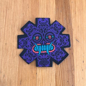 May include: A purple patch with a stylized face design, featuring blue eyes, a turquoise mouth, and pink accents. The design is set against a black outline with a decorative pattern. The text "1993 RED HOT CHILI PEPPERS" is visible.