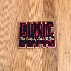 May include: Black and red embroidered patch featuring the name "ELVIS" in bold letters, with the text "The King of Rock & Roll" underneath in gold. The patch is rectangular and has a gold border.