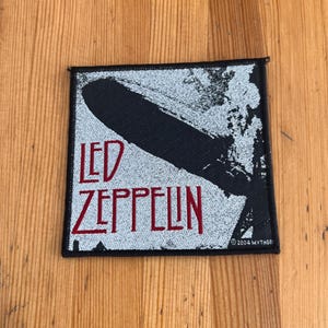 May include: Square patch featuring the Led Zeppelin logo. The design includes a black airship over a silver background, with the band's name in red letters. The patch has a black border and the text "©2004 MYTHGE".