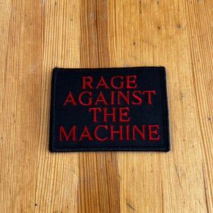 May include: A black rectangular patch with the words "RAGE AGAINST THE MACHINE" embroidered in red. The patch is likely for a jacket, bag, or other apparel item. The patch is on a wooden surface.