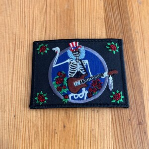 Puede incluir: Un parche rectangular negro con un esqueleto tocando la guitarra dentro de un círculo azul. El esqueleto lleva un sombrero de copa rojo, blanco y azul. Rosas rojas están dispersas alrededor del diseño.