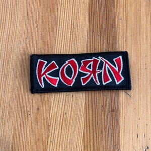 May include: Black and white embroidered patch with the word "Korn" in red lettering.