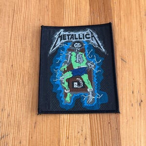 May include: Black square patch featuring the band name "METALLICA" in white, stylized lettering. The patch depicts a green and blue illustration of a skeleton in an electric chair, with blue lightning bolts surrounding it. The patch has a black border.