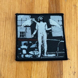 Op de afbeelding: Zwart-witte geborduurde patch met een figuur met vleugels die voor een raam staat. De tekst "Antichrist Superstar" en "©1996 Marilyn Manson/Rock Express" is zichtbaar.