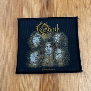 May include: Black Opeth patch featuring the band's logo in gold script above a graphic of five faces. The patch has a black border and the text "©2005 Opeth" at the bottom. The patch is rectangular.