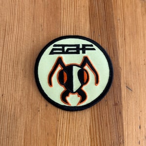 Pode incluir: Um patch circular com uma borda preta, apresentando um fundo verde claro. O patch tem as letras "AAF" em preto no topo. Abaixo das letras está um design estilizado de formiga em preto e laranja.