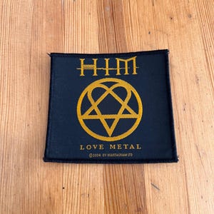 Op de afbeelding: Zwart vierkantige patch met gouden tekst en een heartagram symbool. De tekst luidt "HIM" bovenaan, met "LOVE METAL" en copyright informatie onderaan. Waarschijnlijk een band patch.
