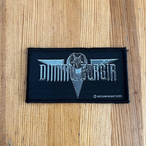 Peut inclure: Écusson rectangulaire noir avec le nom du groupe "Dimmu Borgir" en lettres argentées. L'écusson présente un pentagramme et le texte "NUCLEAR BLAST 2003".