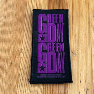 May include: Black rectangular patch with the band name "Green Day" in purple block letters. The text is stacked vertically with stars between the words. The bottom of the patch includes copyright information.