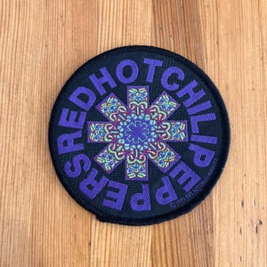 May include: A round black patch with the words "RED HOT CHILI PEPPERS" in purple around the edge. The center features a colorful, symmetrical design in shades of purple, green, and blue.
