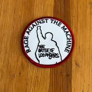 Może przedstawiać: Okrągła naszywka z białym tłem i czerwoną obwódką. Naszywka przedstawia czarną sylwetkę osoby z uniesioną ręką oraz czarny napis "RAGE AGAINST THE MACHINE" i "THE BATTLE OF LOS ANGELES".
