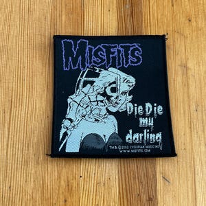 Può includere: Toppa quadrata nera con la scritta "Misfits" in viola, sopra una grafica di scheletro. Il testo "Die Die my darling" è sotto la grafica. La toppa ha un bordo nero ed è su una superficie di legno.