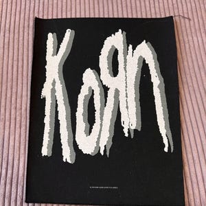 Pode incluir: Patch quadrado preto com o nome da banda "Korn" em uma fonte branca estilizada com um efeito de sombra cinza. O patch é provavelmente feito de tecido e foi projetado para ser costurado ou passado a ferro em roupas ou acessórios.