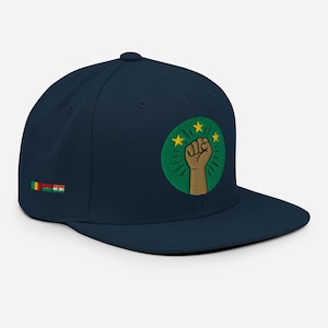 Puede incluir: Gorra de béisbol azul marino con visera plana, con un diseño bordado. El diseño incluye un puño marrón dentro de un círculo verde con estrellas y rayos amarillos. Pequeñas banderas bordadas en el lateral.