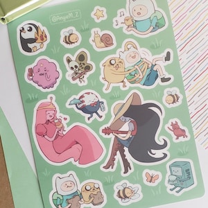 May include: A sheet of Adventure Time stickers featuring various characters like Finn, Jake, Princess Bubblegum, and Marceline. The stickers are colorful and cartoonish, with a green background and the text "Angie M_Z".
