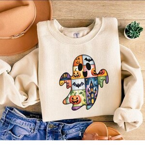 May include: A cream-coloured sweatshirt with a colourful ghost design made up of Halloween-themed patches. The ghost has a patchwork appearance with pumpkins, bats, and a haunted house. The sweatshirt is paired with blue denim shorts and a brown hat.