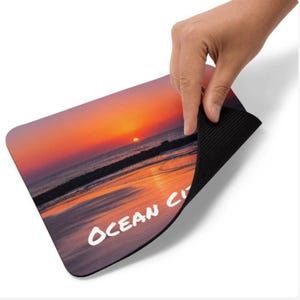 May include: A rectangular mouse pad featuring a vibrant sunset over the ocean with the text OCEAN CITY printed in white. The mouse pad has a smooth top surface with a colorful beach scene and a black, textured, non-slip rubber base underneath.