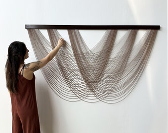 May include: A brown wooden frame holds a wall hanging made of thin, light brown cords woven in a wave pattern.