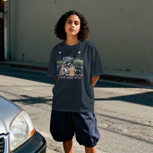 May include: Navy blue t-shirt featuring a raccoon wearing sunglasses and a hat, holding a soccer ball and a drink. The text "I LIVE HERE NOW" is below the graphic. The shirt is paired with navy blue shorts.