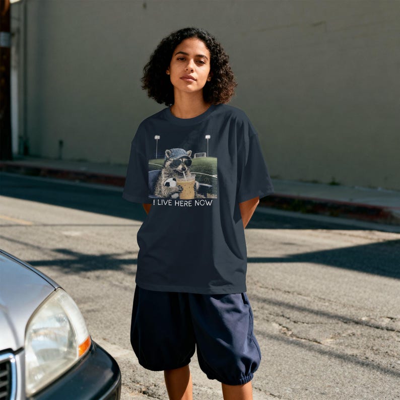 May include: Navy blue t-shirt featuring a raccoon wearing sunglasses and a hat, holding a soccer ball and a drink. The text "I LIVE HERE NOW" is below the graphic. The shirt is paired with navy blue shorts.