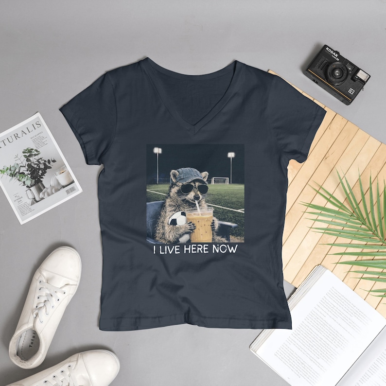 May include: Navy blue V-neck t-shirt featuring a raccoon wearing sunglasses and a hat, holding a soccer ball and a drink. The graphic includes the text "I LIVE HERE NOW". The raccoon is sitting in a stadium.