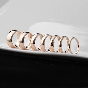 May include: A collection of seven rose gold rings of varying sizes, arranged in a row. The rings have a smooth, polished finish and a rounded profile. The background is a gradient of white and black.