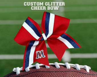 May include: A red, white, and blue cheer bow with glitter accents and the letter "M" on a football. The bow has a custom glitter design and is set against a green background with white lines. Text at the top reads "CUSTOM GLITTER CHEER BOW."