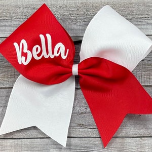 May include: A red and white cheer bow with the name "Bella" written in white on the red bow.