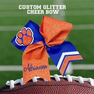 May include: A custom glitter cheer bow in blue and orange, with the name Adrianna embroidered on one tail. The bow features a paw print and chevron design, set against a football field background.