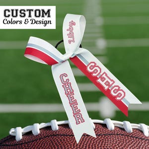Puede incluir: Una cinta blanca para el pelo con detalles en rojo y gris y el texto "Libby" en rojo. La cinta est&aacute; sujeta a un bal&oacute;n de f&uacute;tbol y tiene el texto "SHS" y "CHEER" en rojo. La imagen tambi&eacute;n incluye el texto "CUSTOM Colors & Design".