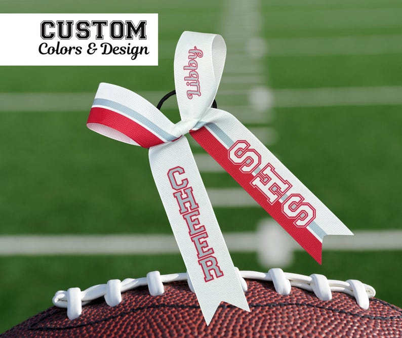 Puede incluir: Una cinta blanca para el pelo con detalles en rojo y gris y el texto "Libby" en rojo. La cinta est&aacute; sujeta a un bal&oacute;n de f&uacute;tbol y tiene el texto "SHS" y "CHEER" en rojo. La imagen tambi&eacute;n incluye el texto "CUSTOM Colors & Design".
