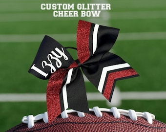 May include: A custom glitter cheer bow in black, red glitter, and white. The bow is personalized with the name "Izzy" in white script. The bow is resting on a football.