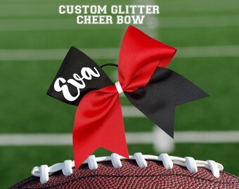 May include: A custom glitter cheer bow in red and black, with the name "Eva" in white script. The bow is set against a football and a green field background. Text at the top reads "CUSTOM GLITTER CHEER BOW."