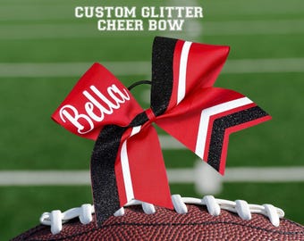 May include: A red and black cheer bow with the name "Bella" in white script. The bow has black glitter accents and white stripes. Text at the top reads "CUSTOM GLITTER CHEER BOW". The bow is resting on a football.