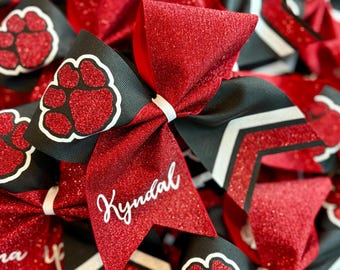May include: Red glitter cheerleading bows with black and white accents. One bow features the name "Kyndal" in white script. Other bows have a paw print design.