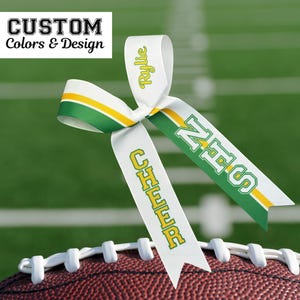 Puede incluir: Un lazo de animadora de cinta blanca con detalles en verde y amarillo, apoyado en un bal&oacute;n de f&uacute;tbol. El lazo tiene el texto "Rylie", "NHS" y "CHEER" en verde y amarillo. El texto "CUSTOM Colors & Design" est&aacute; en la esquina superior izquierda.