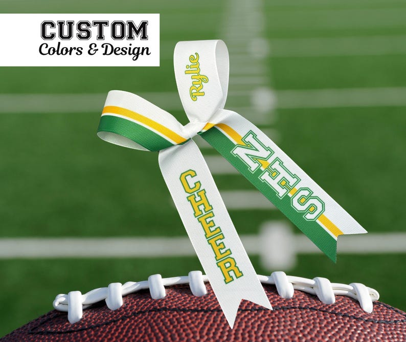 Puede incluir: Un lazo de animadora de cinta blanca con detalles en verde y amarillo, apoyado en un bal&oacute;n de f&uacute;tbol. El lazo tiene el texto "Rylie", "NHS" y "CHEER" en verde y amarillo. El texto "CUSTOM Colors & Design" est&aacute; en la esquina superior izquierda.