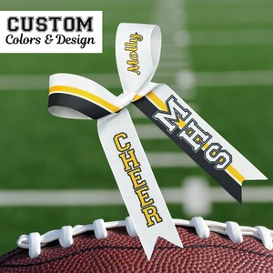 Puede incluir: Un lazo de cinta blanca con detalles en negro y amarillo, colocado sobre un bal&oacute;n de f&uacute;tbol. La cinta muestra los textos "Molly", "MHS" y "CHEER" en amarillo y negro. La esquina superior izquierda tiene el texto "CUSTOM Colors & Design".