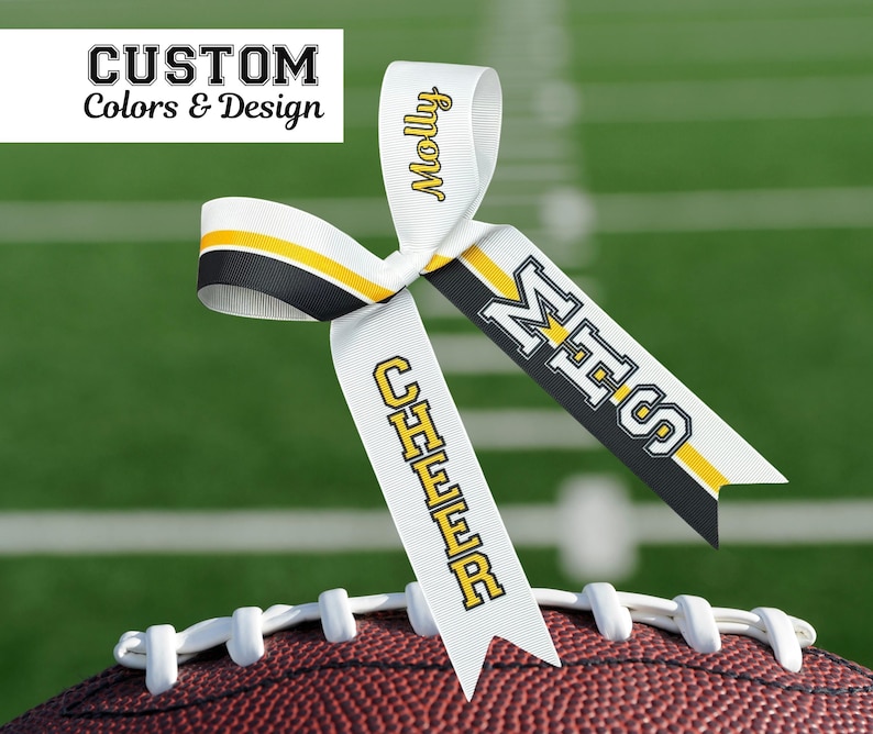 Puede incluir: Un lazo de cinta blanca con detalles en negro y amarillo, colocado sobre un bal&oacute;n de f&uacute;tbol. La cinta muestra los textos "Molly", "MHS" y "CHEER" en amarillo y negro. La esquina superior izquierda tiene el texto "CUSTOM Colors & Design".